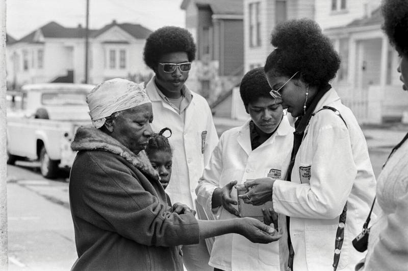 a black and white photo of five African-American people, some in lab coats, standing outside doing a blood draw on one of them, an older woman