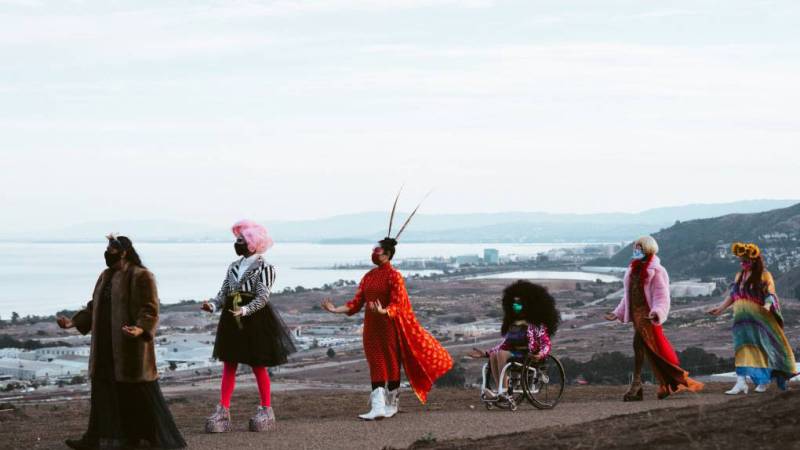 Artists in a processional wearing colorful clothing atop a coastline ridge, with the sea in the background.