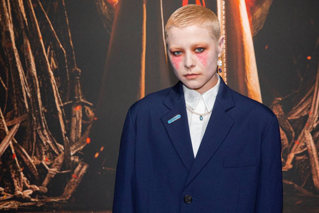 An androgynous white person with short slicked back blonde hair and dramatic pink makeup stands on a red carpet wearing an oversized blue suit jacket.
