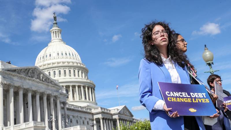 Woman in blue suit holds sign saying "cancel deb, change lives" with capitol in background