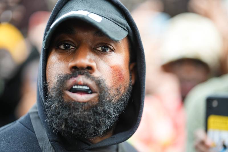 A bearded Black man wearing a black hoodie and baseball cap stares into the camera, mouth slightly open. He is wearing a mouthguard with Balenciaga written on it.