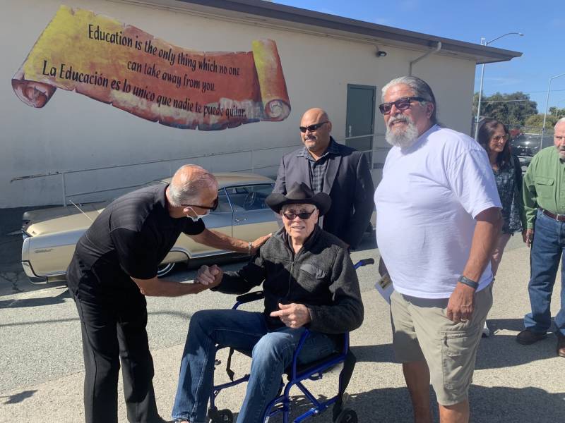 four men pose for a photo outside a school, with one in a black cowboy hat in a wheelchair