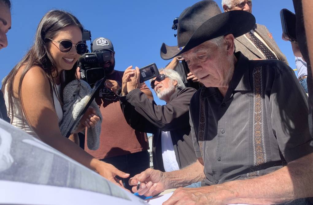 an older man in a black cowboy hat signs autographs for a crowd while photographers snap photos
