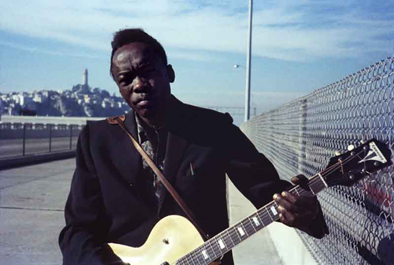 An older black man plays a guitar on a pier with Coit Tower in the background