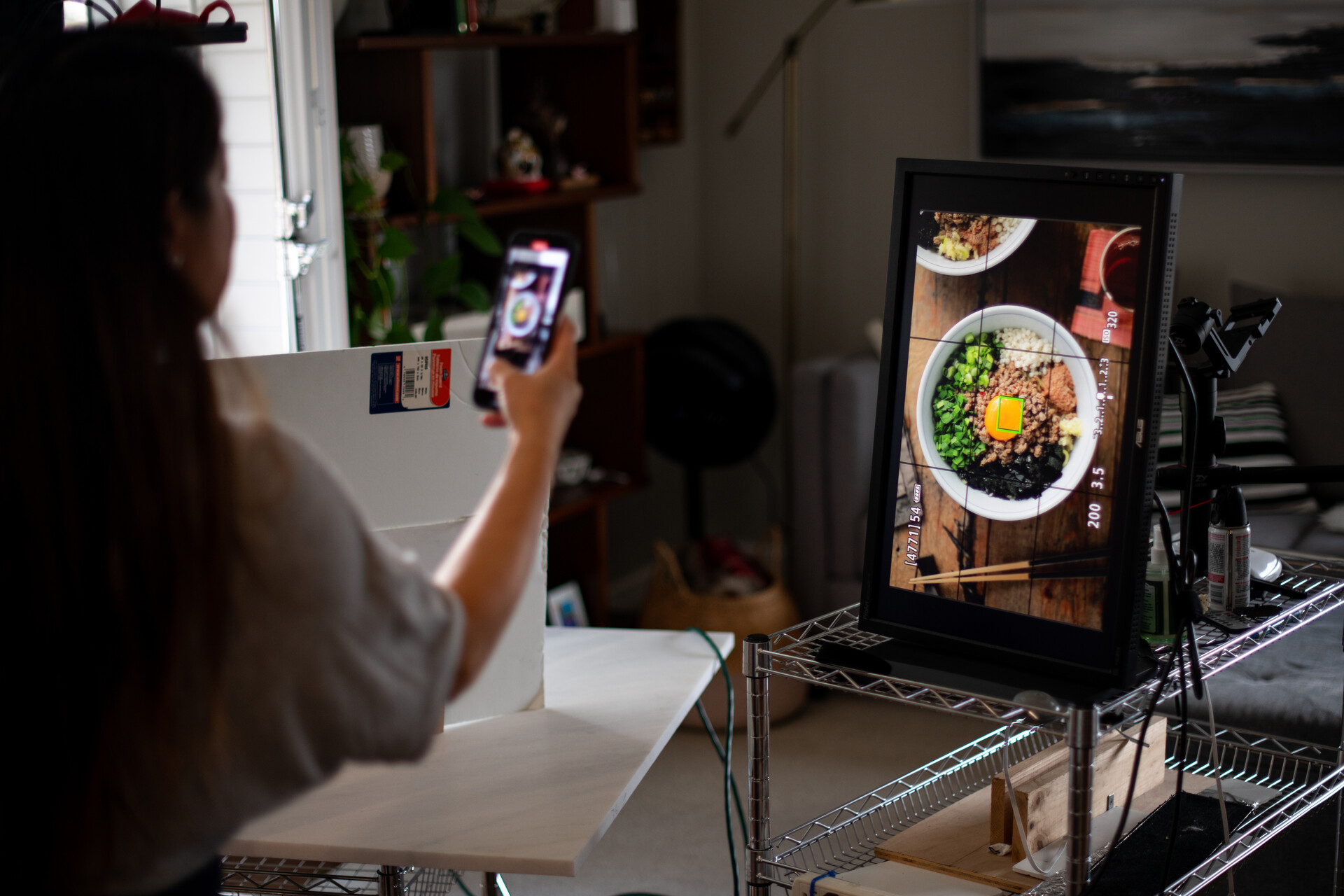A woman uses her cellphone to take a photo of a picture of a bowl of noodles that's projected onto a large flat-screen monitor.