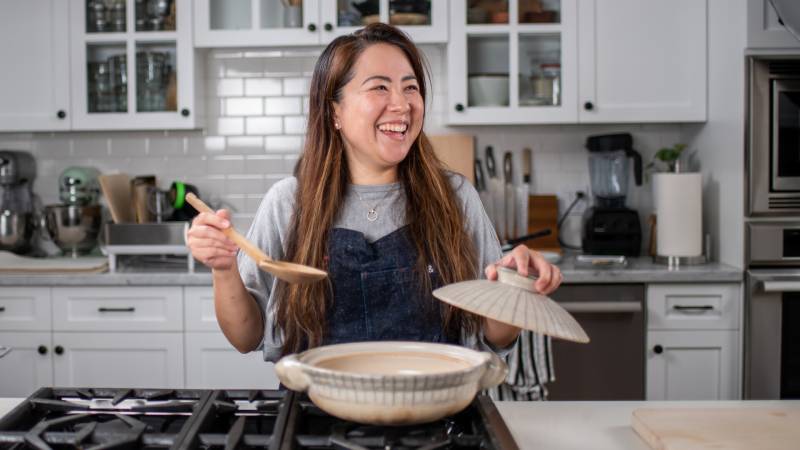 An Asian woman in a dark blue apron smiles while standing at the stove with a ladle and a pot lid in her hands.