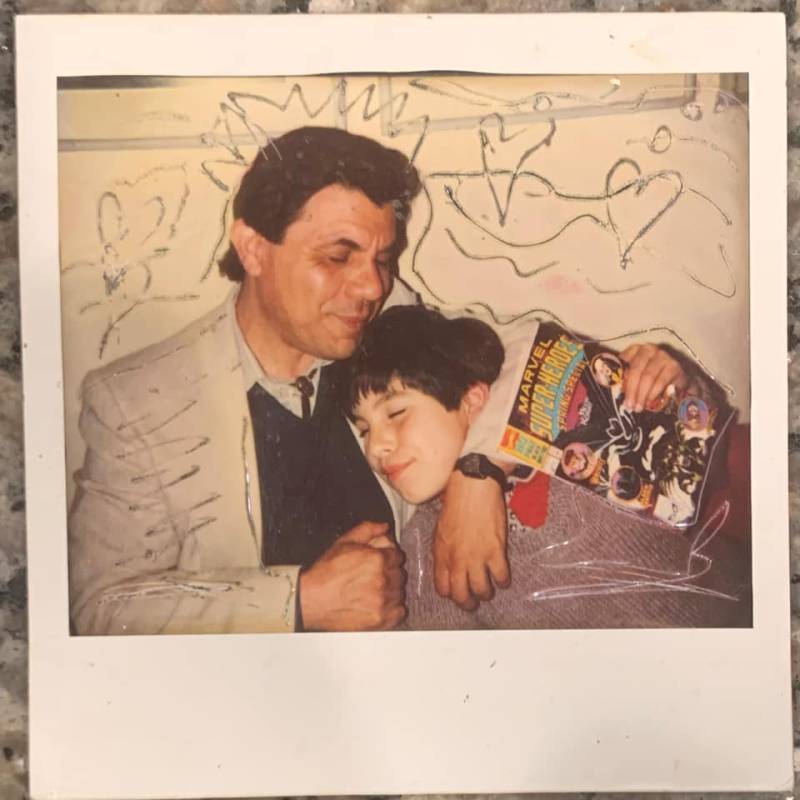 A polaroid photo of young Rio Yañez holding a comic book and embraced by his father Rene.