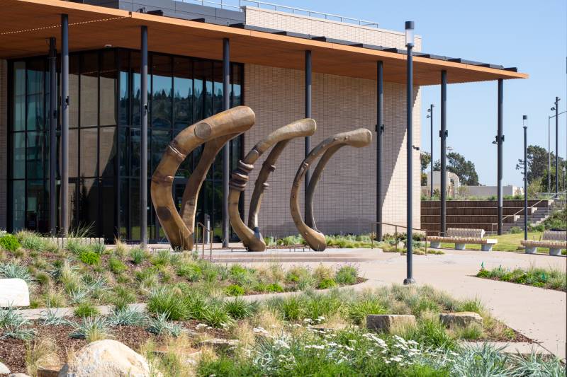 View of building exterior with tall shaded awning and three-part bronze sculpture in foreground with plantings