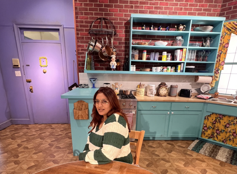 A woman sits at a kitchen table on a set resembling one from 1990s TV show, 'Friends.'