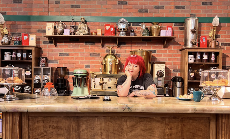 A woman with bright red hair stands behind a coffee shop counter, head resting on chin.