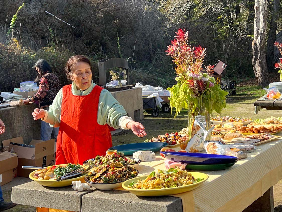 An elderly woman in a red apron is preparing a large meal for a family gathering outdoors.