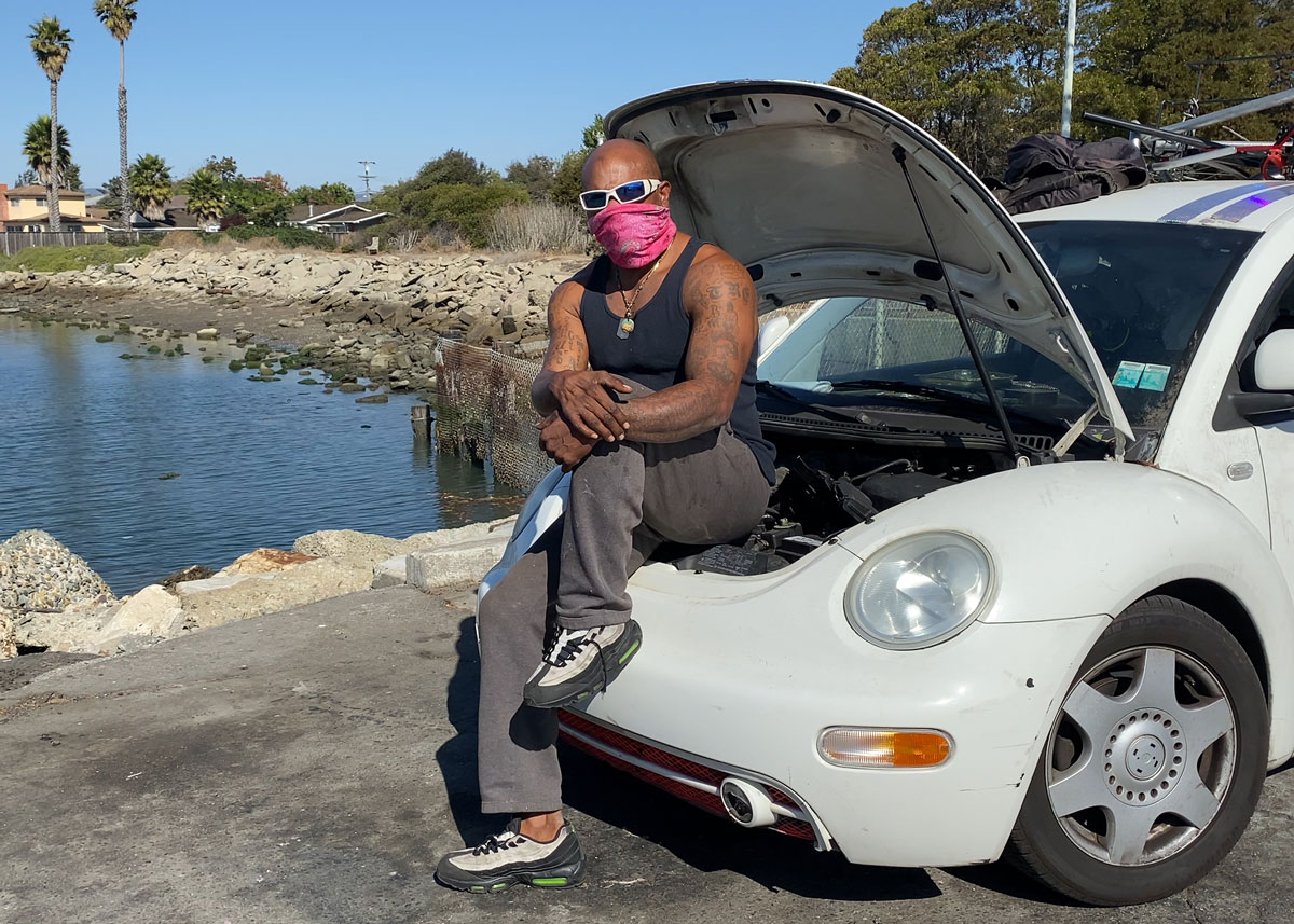 A Black man wearing a red bandana over his nose and mouth poses on the engine of a VW Bug with the hood up