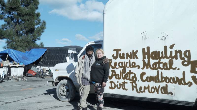 Man and woman stand in front of white truck with hand-written "hauling" advertising on side