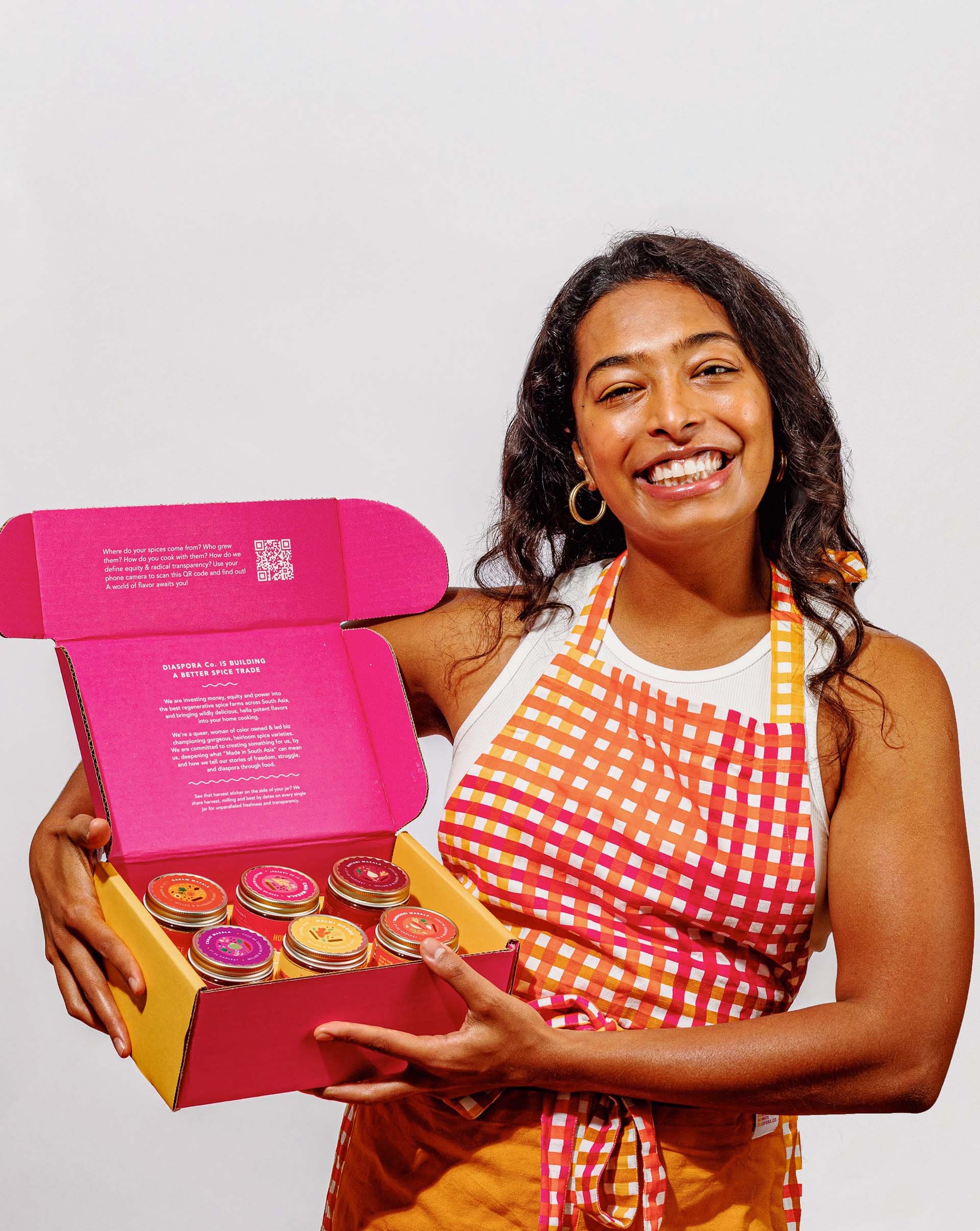 A smiling woman in a checkered pink and yellow apron shows off a box of jarred spices in a matching pink and yellow box. 