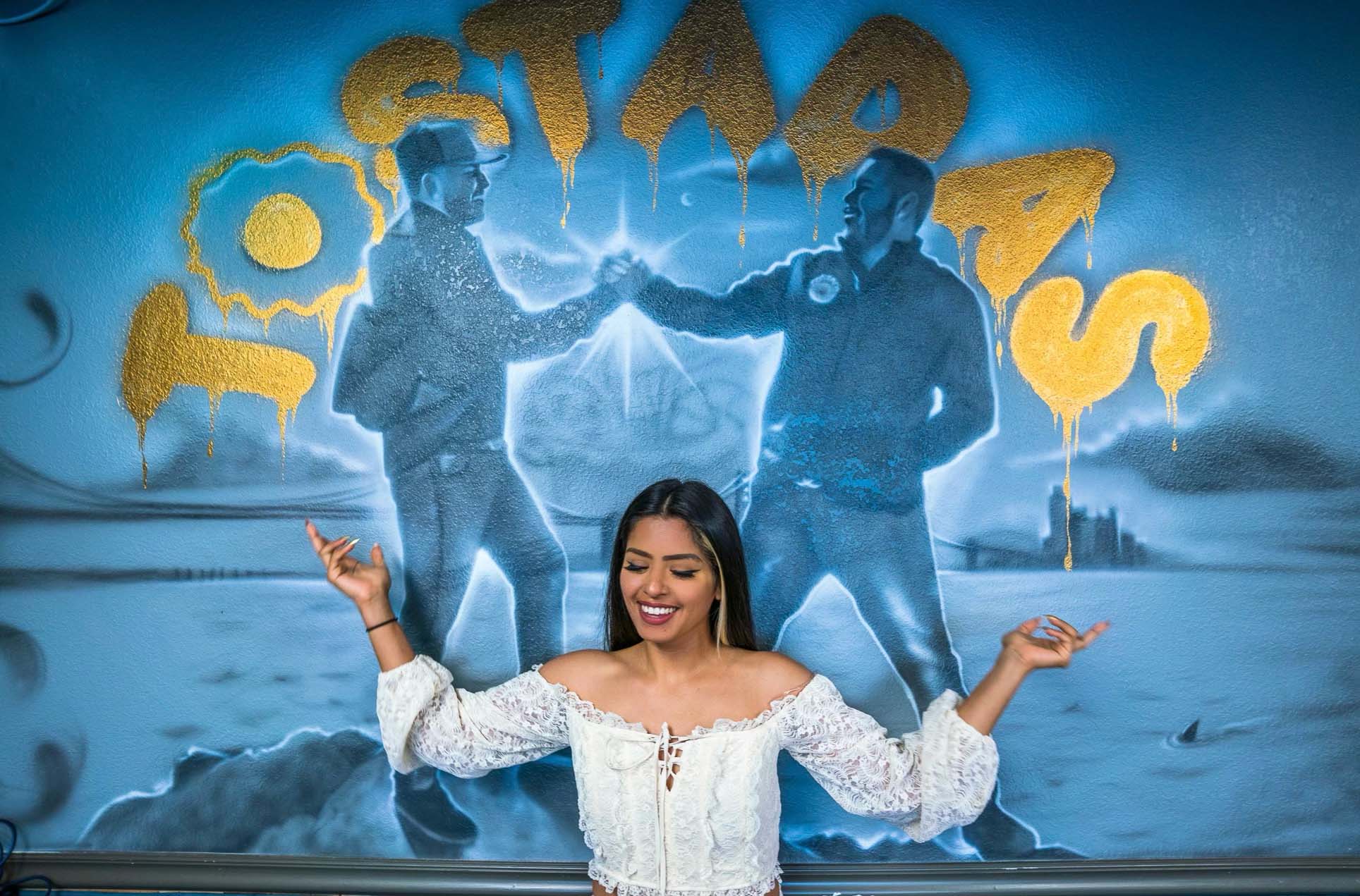 A young Latina stands in front of a mural painted inside a restaurant, depicting two undocumented Latino brothers shaking hands