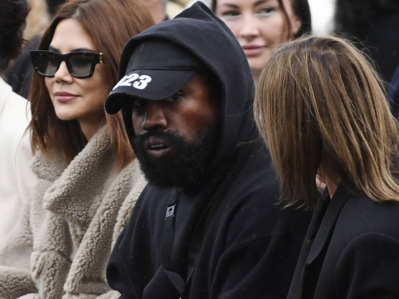 A bearded Black man wearing a black hoodie and black baseball cap sits in a row of seats with white women sitting either side of him.