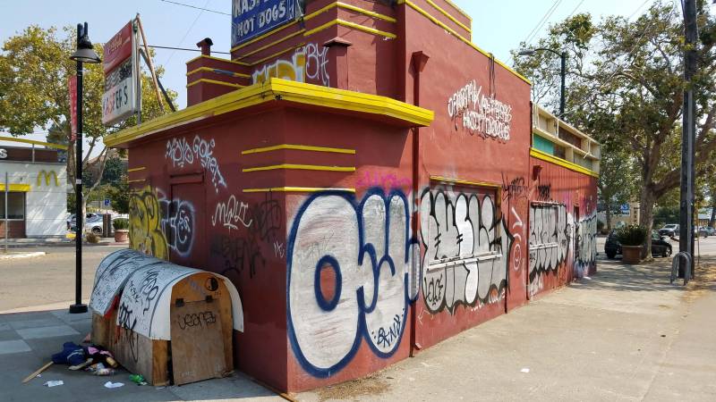 A side profile of Kasper's, a local hot dog business, with a McDonalds in the background. Graffiti cover the sides, and it appears that a homeless person is living on one side of the walls.
