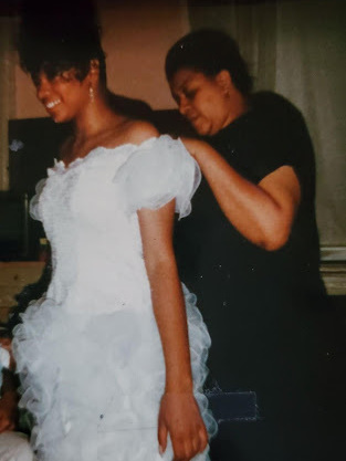 A middle-aged Black woman helps a younger smiling Black woman put on a white prom-style dress.