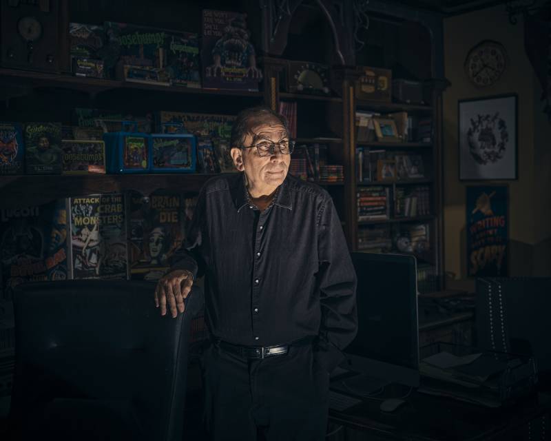 A senior bespectacled man, dressed all in black, stands in a darkened room before an expansive bookcase. His figure is lit from underneath.