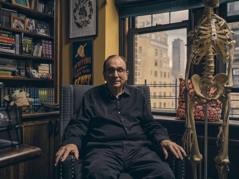 A senior gentleman, dressed in all black and wearing spectacles, sits in an armchair next to a life-size skeleton. Behind him are packed bookshelves and a window.