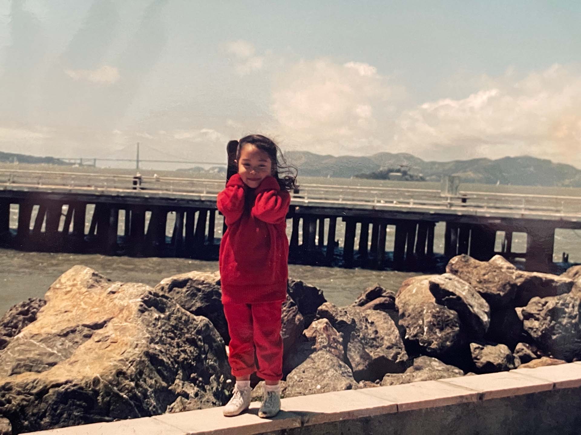 A young girl with windswept hair dressed in all red; the pier and waterfront at Treasure Island is in the background.