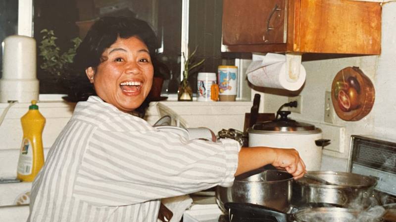 A woman looking up at the camera and smiling with delight as she tends to a pot on the stove.