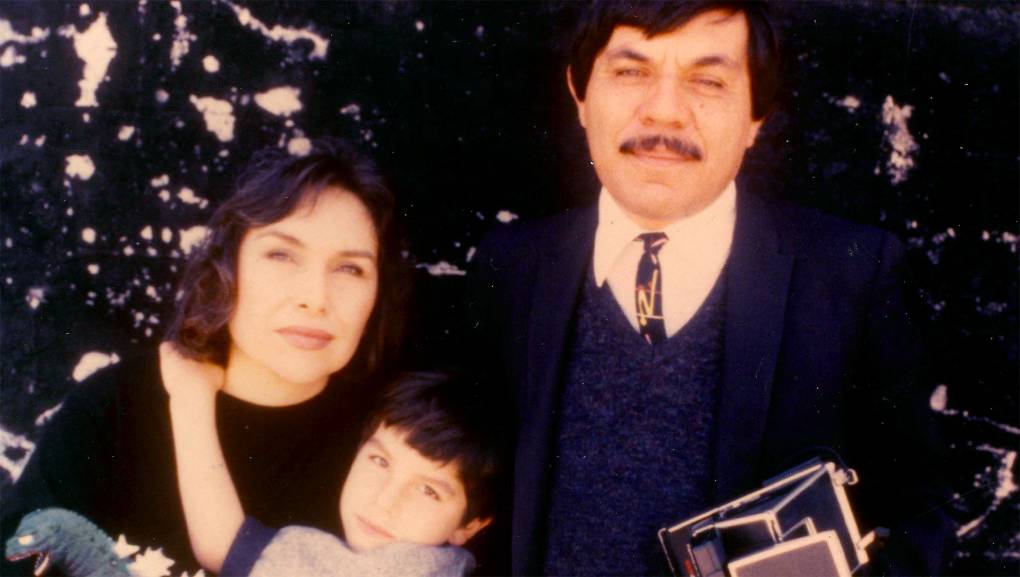 A family photo of a young Rio Yañez in the center of his parents. His mother, on the left, holds Rio's toy Godzilla as he hugs her. His father, on the right, holding a vintage camera.