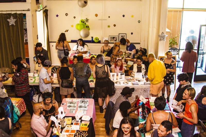 A crowd of young queer people browses through zines displayed at tables.