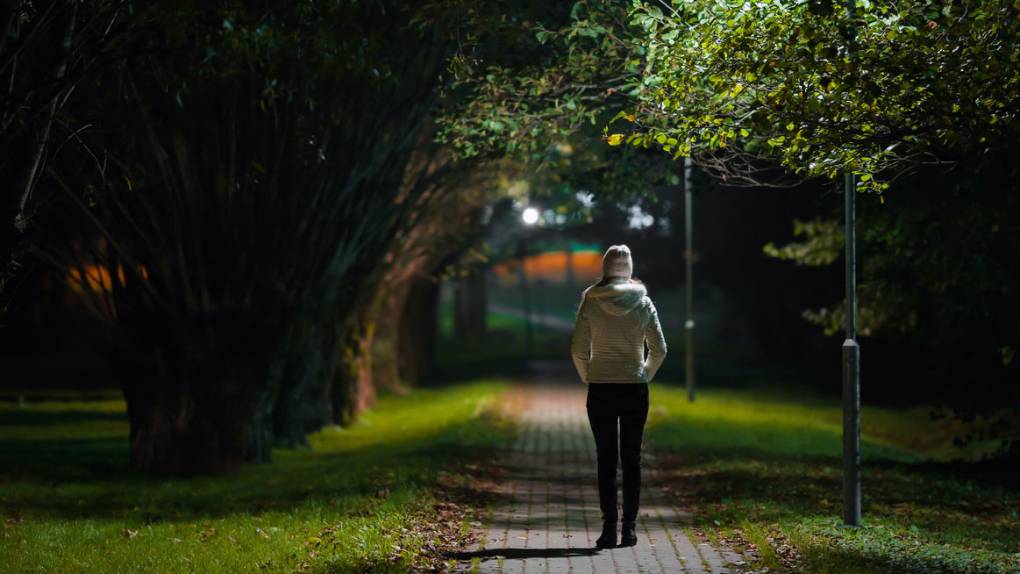 A young woman, back to the camera, walks alone at night, illuminated by a lamp along a park path