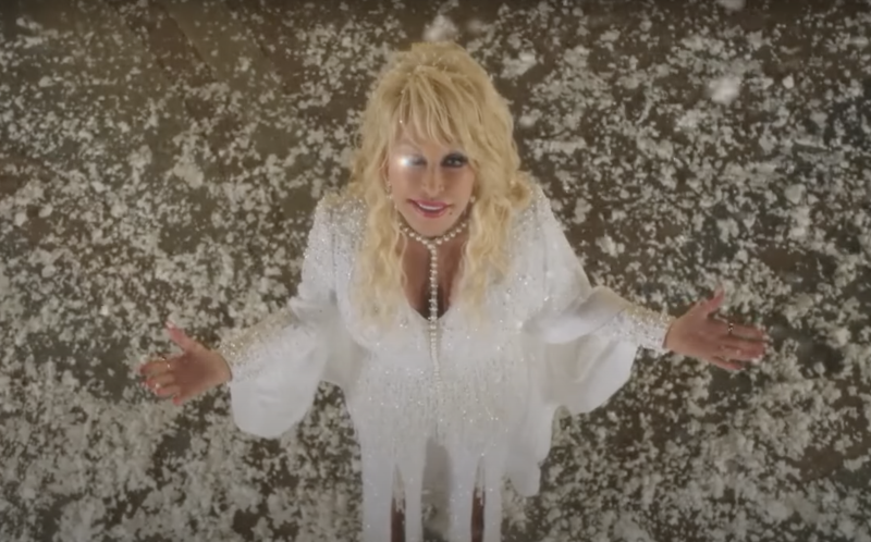 Dolly Parton stands wearing a white dress, surrounded by falling snow, gazing upwards towards the camera. She is winking, a bright light covering her closed eyelid.