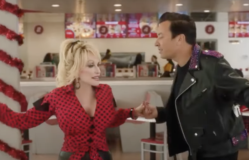 Dolly Parton in a tight red and black that's tied at the waist. Jimmy Fallon in a leather jacket. They are dancing inside a diner. 