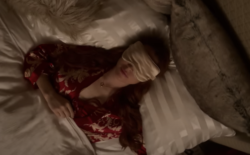 A woman lying on silky white pillows, under a duvet, wearing a red nightgown and a white eye mask.