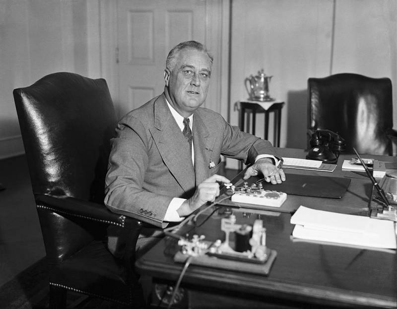 A distinguished older man sits at his desk about to press down on a device that resembles a morse code machine.