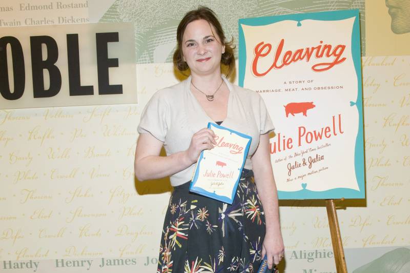 A woman dressed casually smiles while holding up a book. A sign behind her reads 'Cleaving, Julie Powell.'