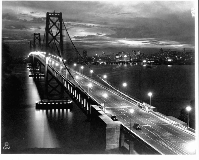 A black and white image of the Oakland-San Francisco Bay Bridge, taken at dusk.