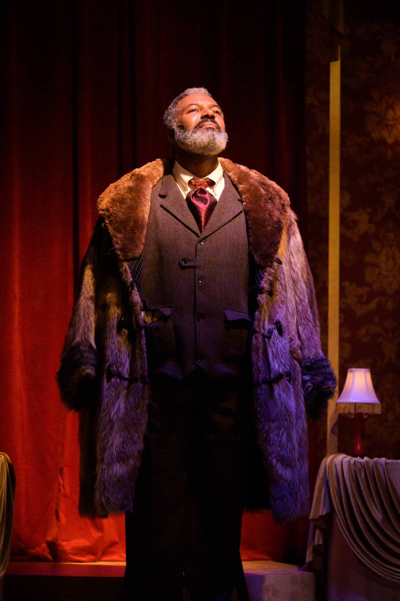 Albert Hodge, a Black Man in a furcoat and a brown suit and red satin tie looks into the distance, a red curtain behind him.