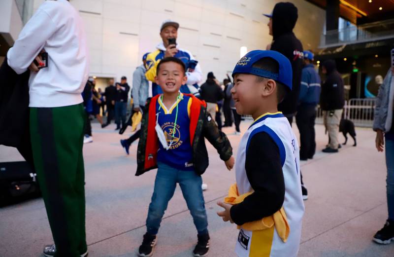 Two small children in Warriors gear laugh with their mother nearby in a crowd of sports fans