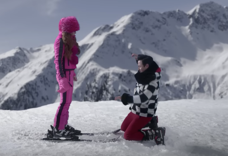 A woman in a fluorescent pink ski suit with black details stands on skis on a mountaintop. A man in a black and white checkered jacket knees before her.