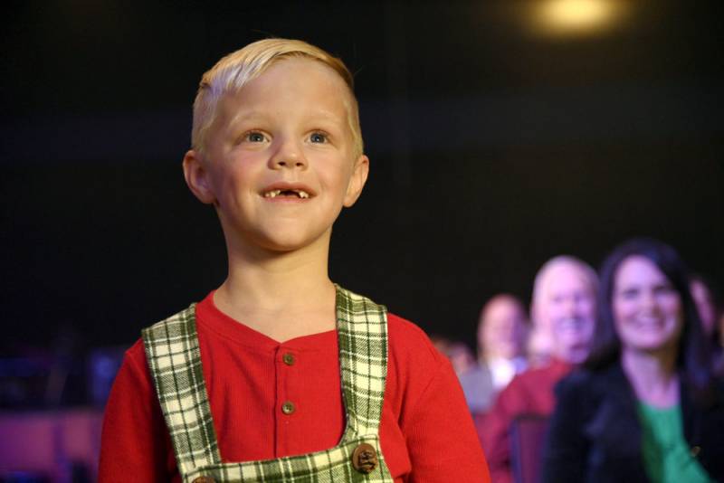 A cute 6 or 7 year old blonde boy, looks off into the distance smiling. He is wearing a red sweatshirt and white and green checked overalls.