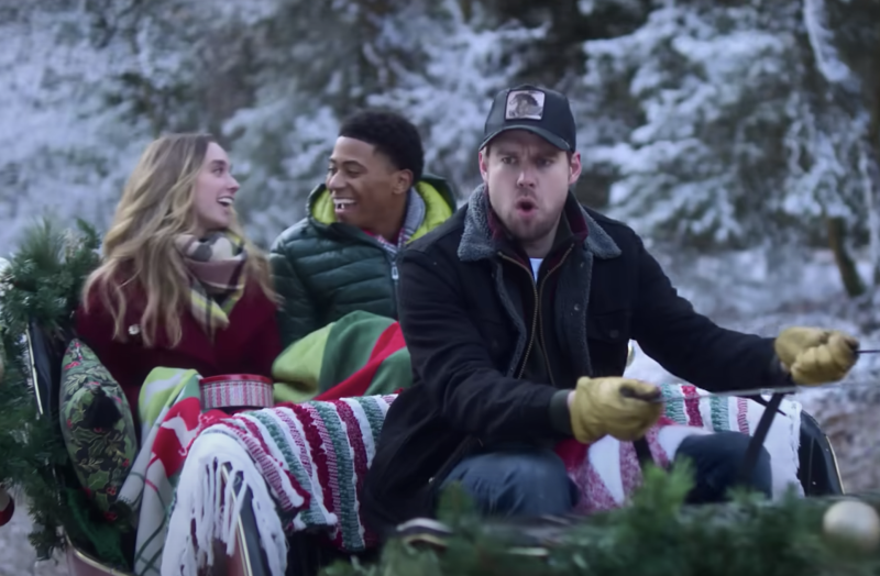 A man driving a sleigh looks into the distance, shocked, while a white blond woman and a handsome Black man gaze at each other adoringly in the back of the sleigh.