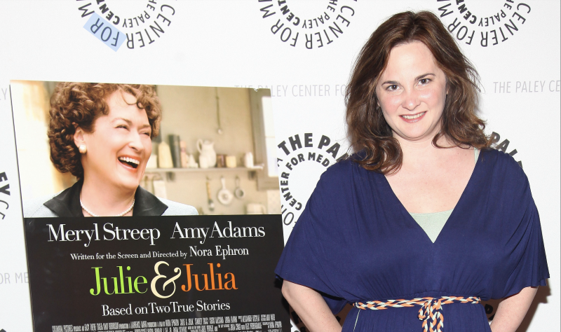 A smiling white woman with shoulder length brown hair stands next to a movie poster for 'Julie & Julia'. She is wearing a casual blue dress and minimal make-up.