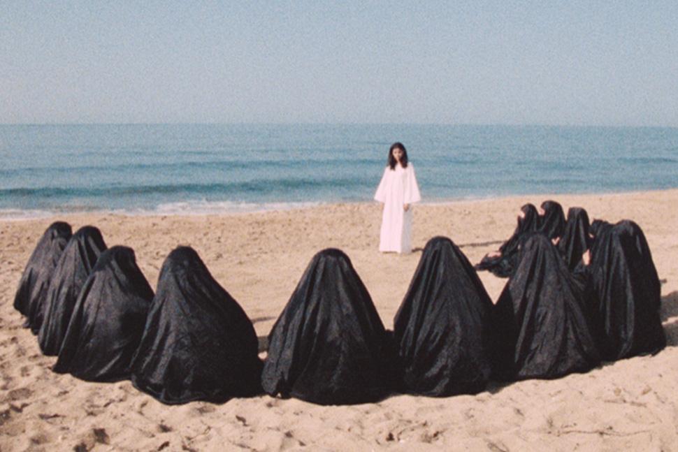 Woman in white dress faces a group of women seated in black burqas on beach