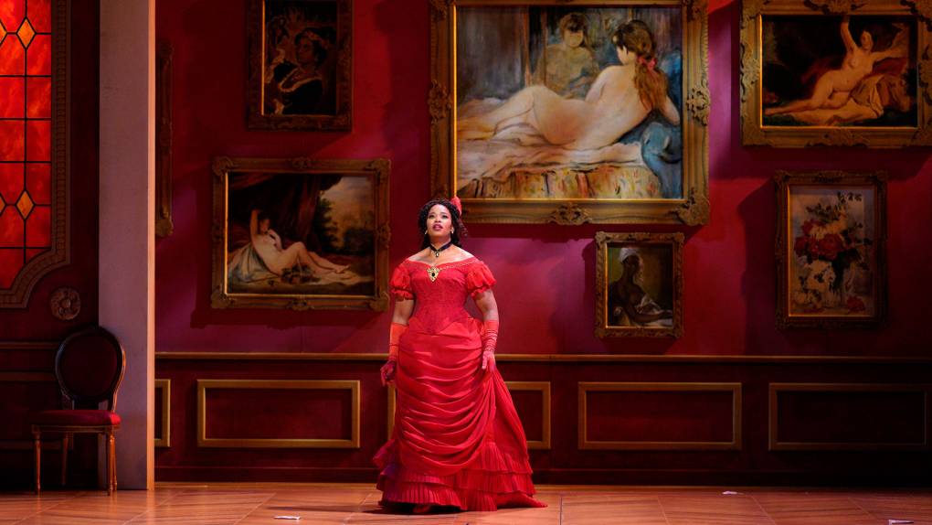 A Black woman in a red dress stands against a parlor wall of deep red, adorned with paintings