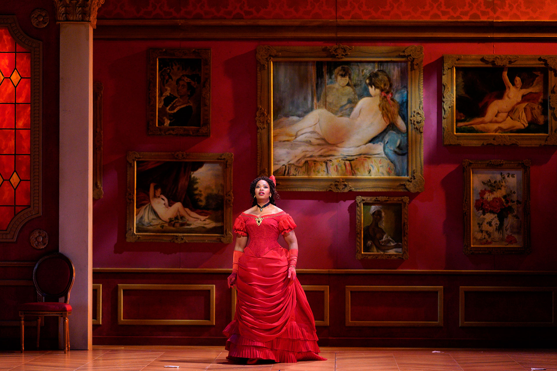 A Black woman in a red dress stands against a parlor wall of deep red, adorned with paintings