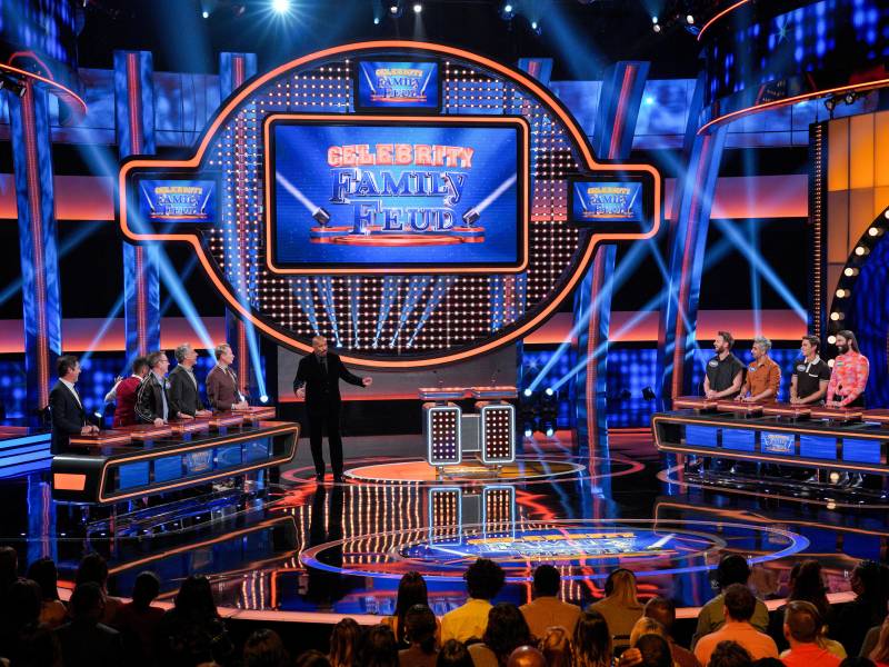 The colorful, neon lit set of 'Family Feud' where two long podiums of people face off underneath a big screen.