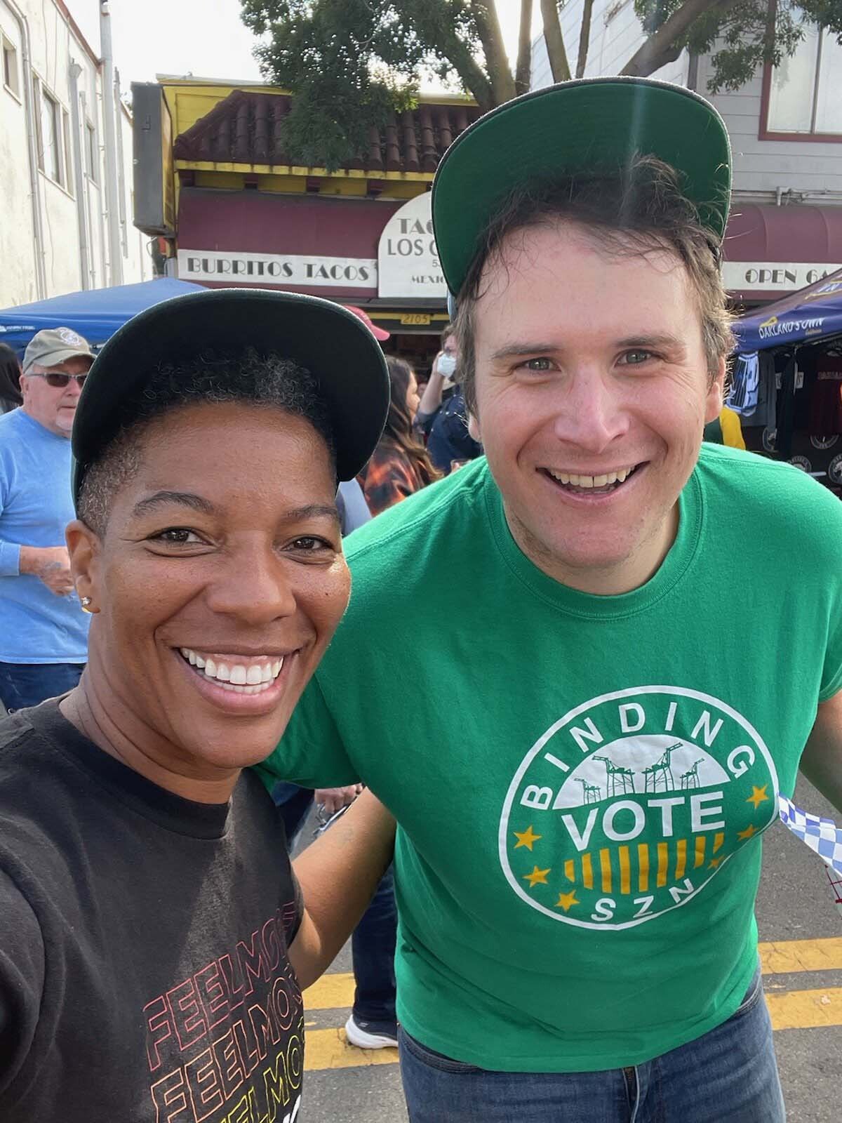 Man in a green "Binding Vote Szn" t-shirt poses with a woman in a black baseball cap.