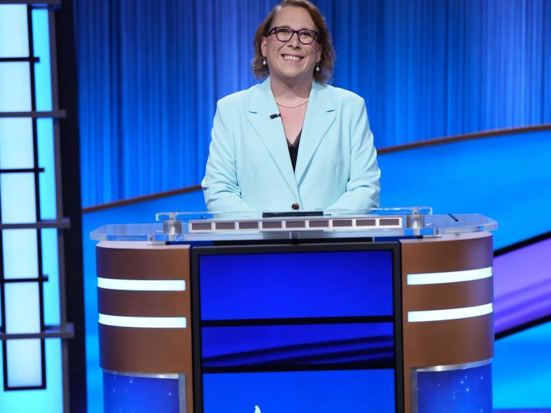 A bespectacled woman in powder blue suit jacket stands behind a game show podium.