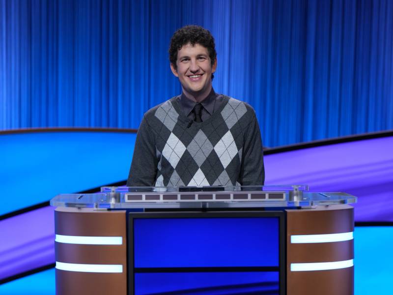 A young man with tight curly black hair, wearing an argyle sweater stands behind a game show podium.