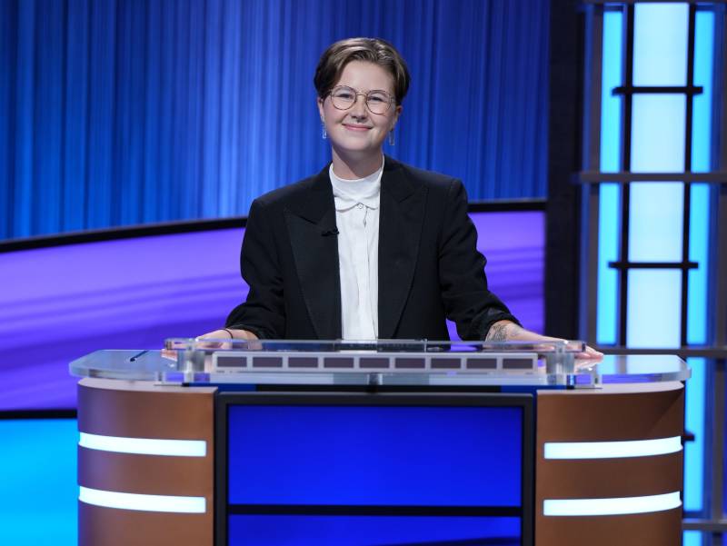 A young woman with short hair and glasses stands behind a game show podium wearing a white shirt and black suit jacket.