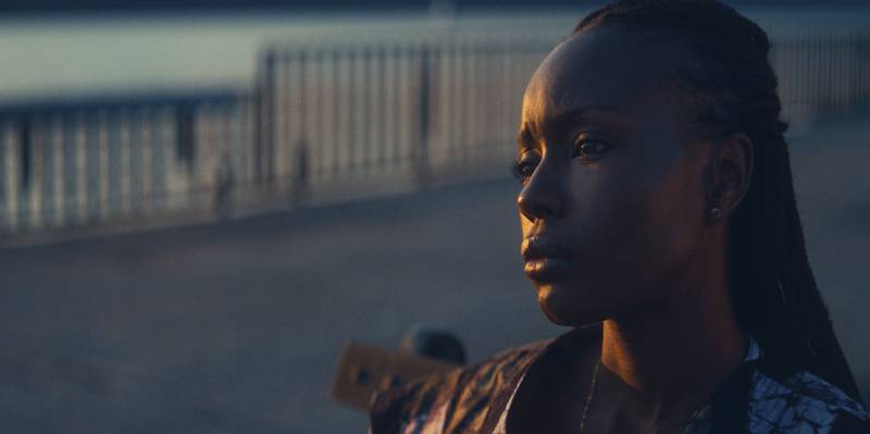 A sullen Black woman looks out over a body of water from an urban environment, as the sun sets.
